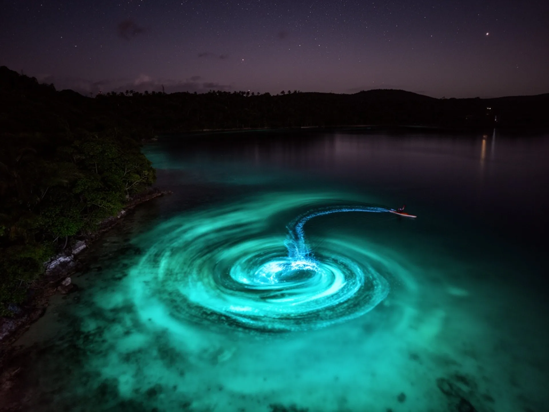 An aerial view of a bioluminescent bay at night in Puerto Rico, glowing turquoise water swirling with each wave, a kayaker leaving a trail of blue-green light, dark jungle coastline framing the scene, stars reflected on the calm water surface, long exposure capturing the ethereal movement of light