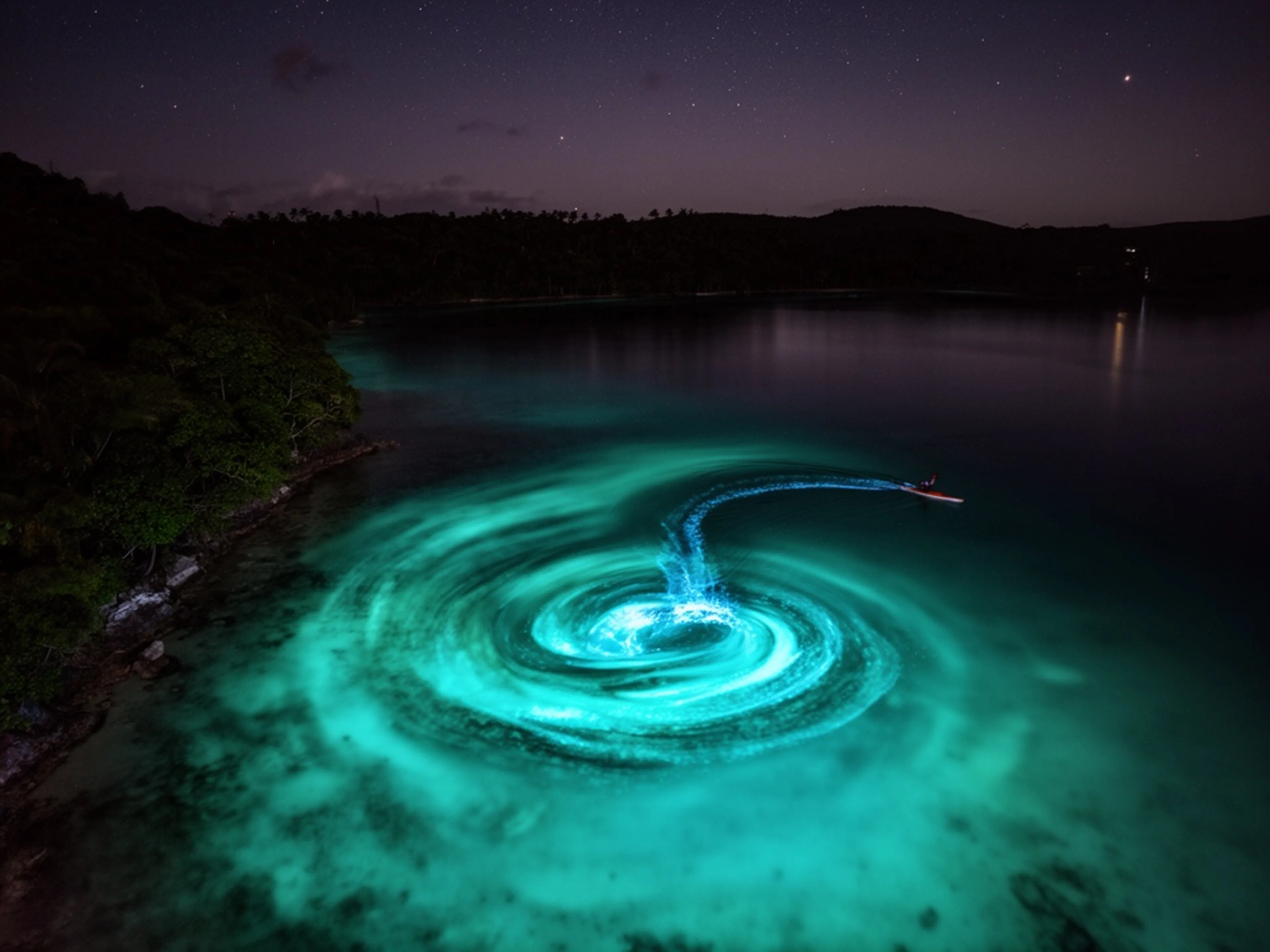 An aerial view of a bioluminescent bay at night in Puerto Rico, glowing turquoise water swirling with each wave, a kayaker leaving a trail of blue-green light, dark jungle coastline framing the scene, stars reflected on the calm water surface, long exposure capturing the ethereal movement of light