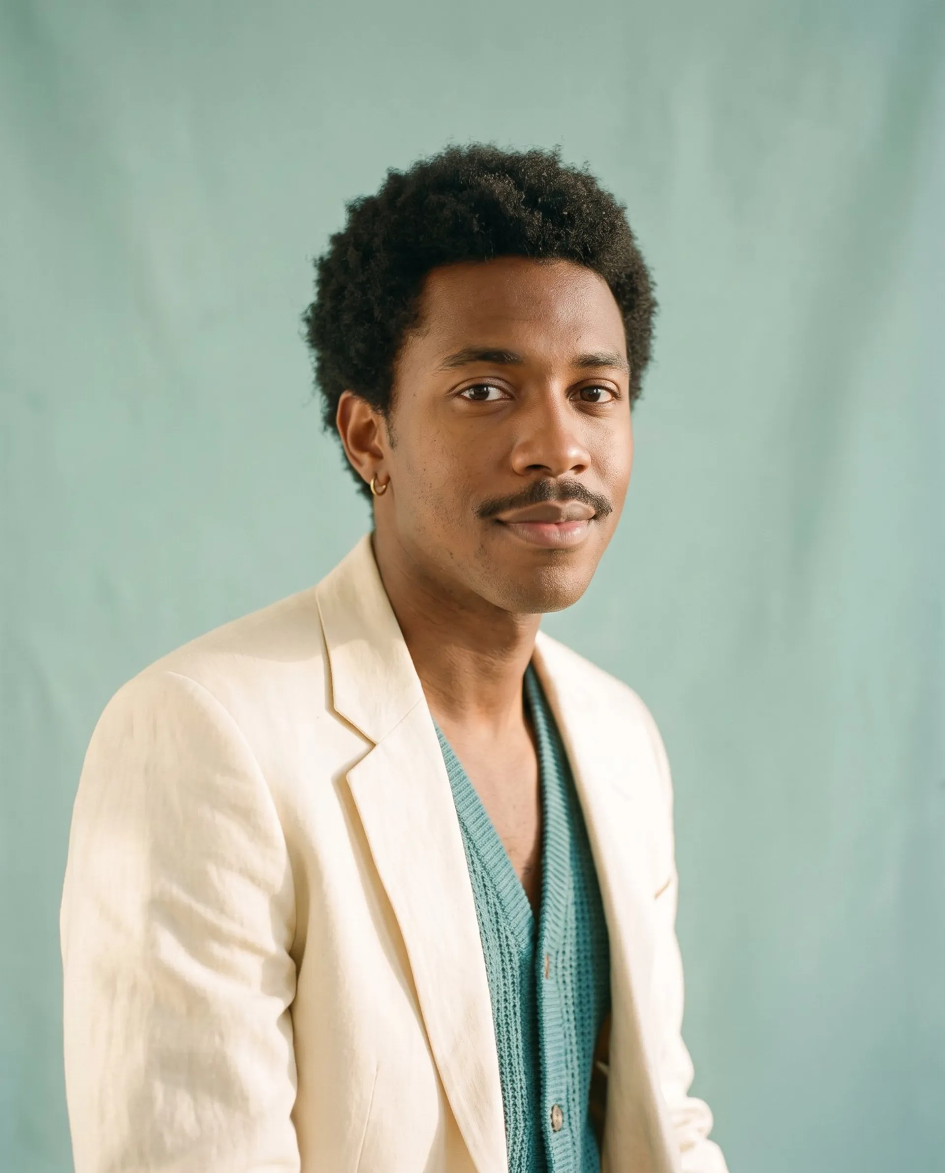 AI-generated studio portrait of a young man with a short afro, a thin moustache and a gold hoop earring, in a cream linen jacket and teal vest